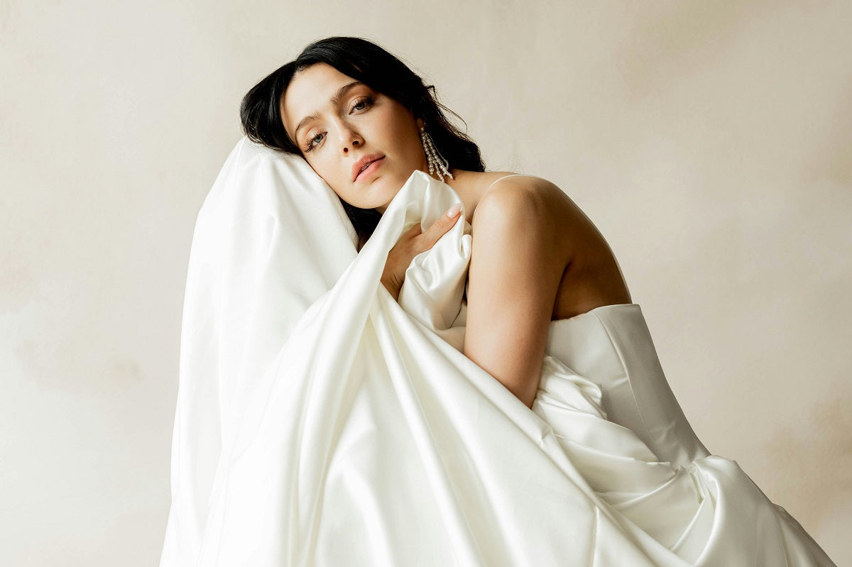 Bride in an ivory satin gown wrapped in its skirt, wearing crystal drop earrings
