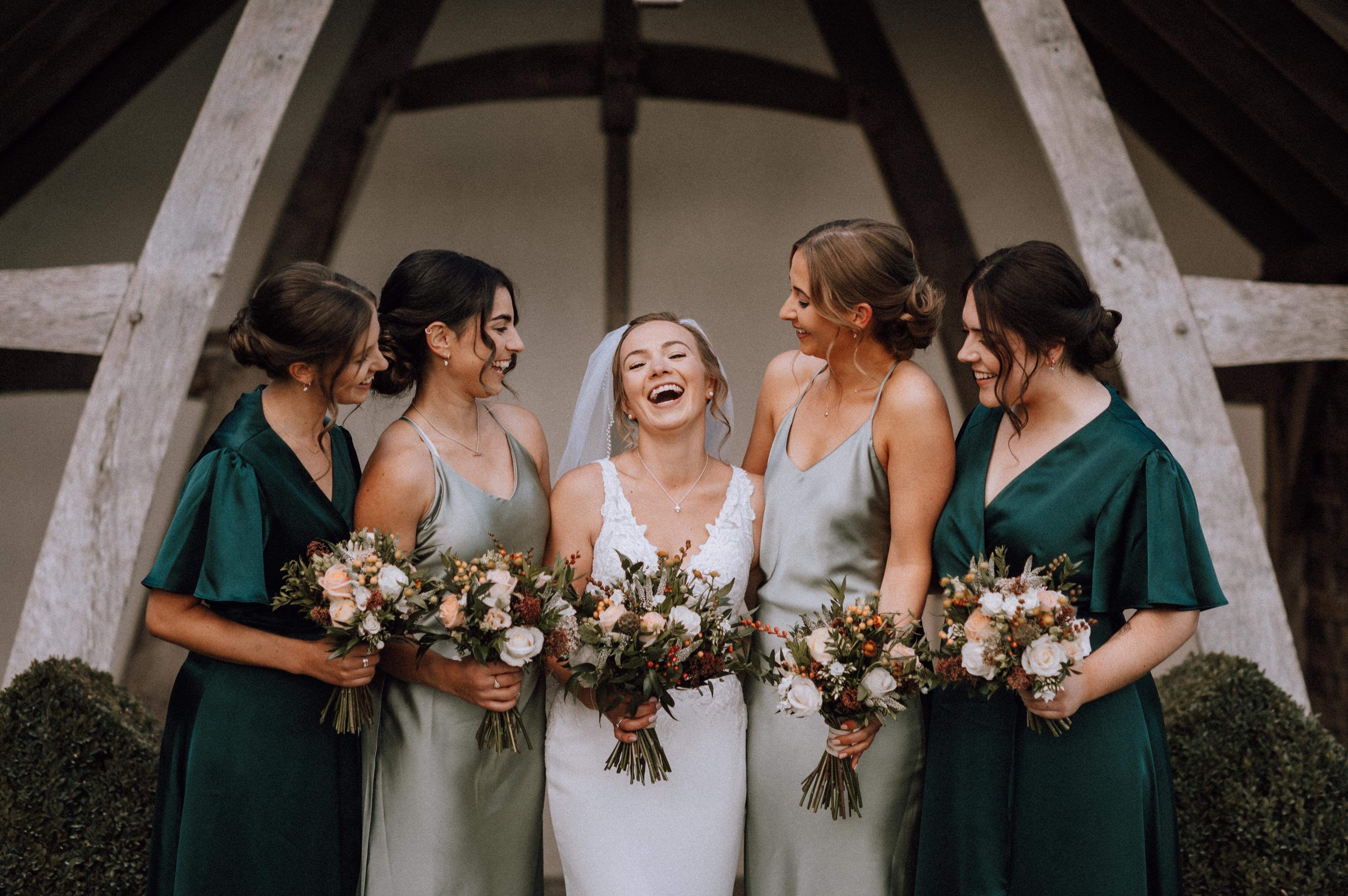 Laughing bride with four bridesmaids in emerald and sage satin dresses at a barn