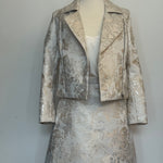 Elegant women's two-piece jacquard outfit on a dress mannequin, featuring a light cream and silver floral-patterned cropped jacket and matching A-line skirt, styled with a soft satin camisole underneath against a plain background.