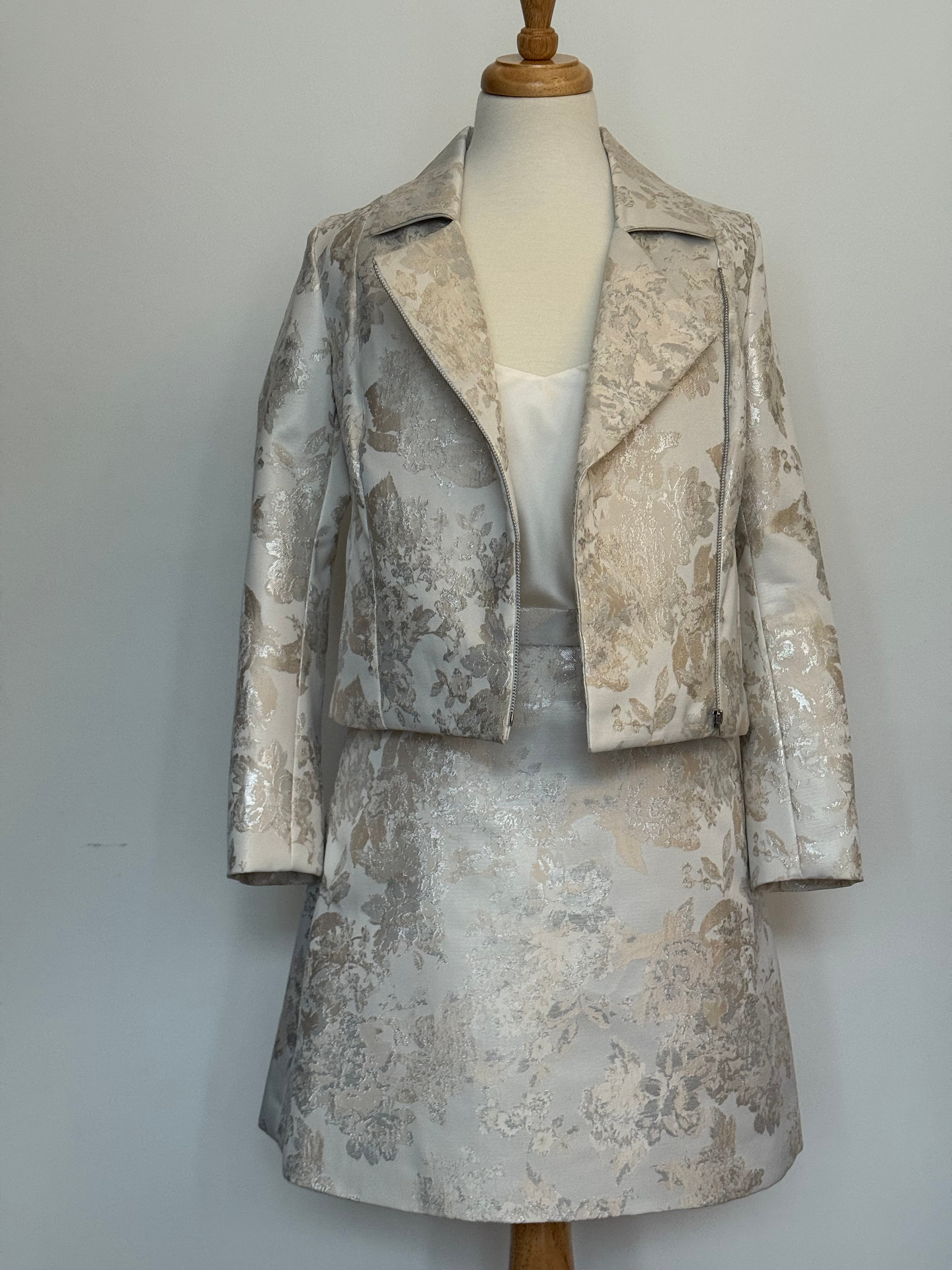 Elegant women's two-piece jacquard outfit on a dress mannequin, featuring a light cream and silver floral-patterned cropped jacket and matching A-line skirt, styled with a soft satin camisole underneath against a plain background.