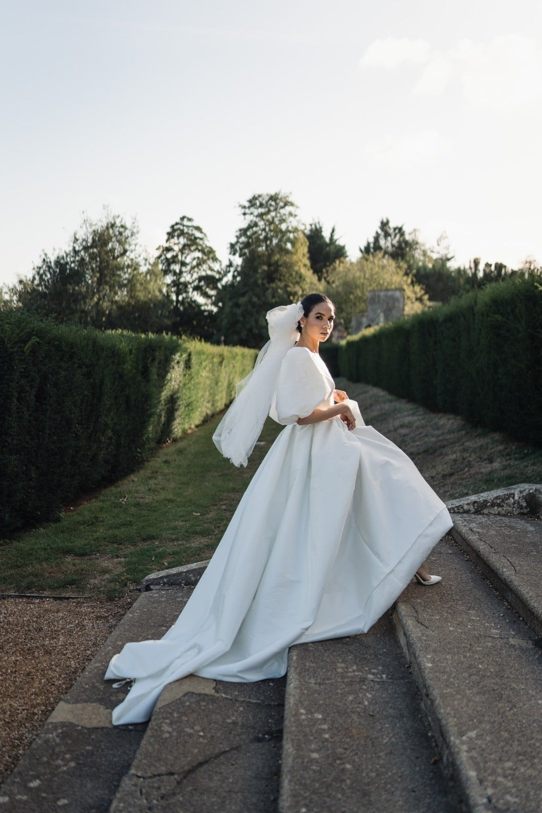 A Model wearing an ivory-colored, an Italian soft tulle bow-shaped veil that extends to her fingertip, standing outdoors.
