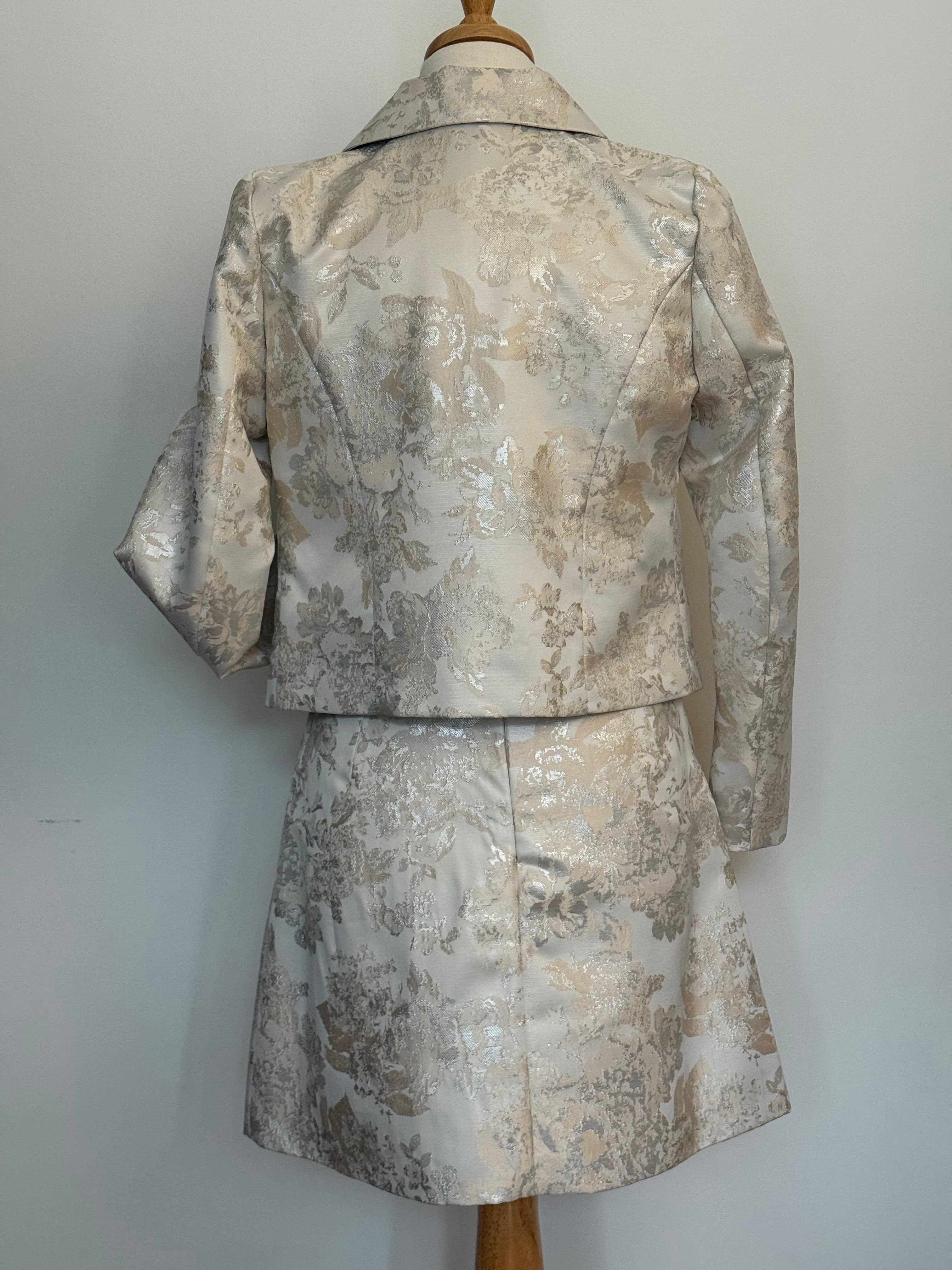 Back of an elegant women's two-piece jacquard outfit on a dress mannequin, featuring a light cream and silver floral-patterned 