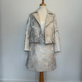 Elegant women's two-piece jacquard outfit on a dress mannequin, featuring a light cream and silver floral-patterned cropped jacket and matching A-line skirt, styled with a soft satin camisole underneath against a plain background.