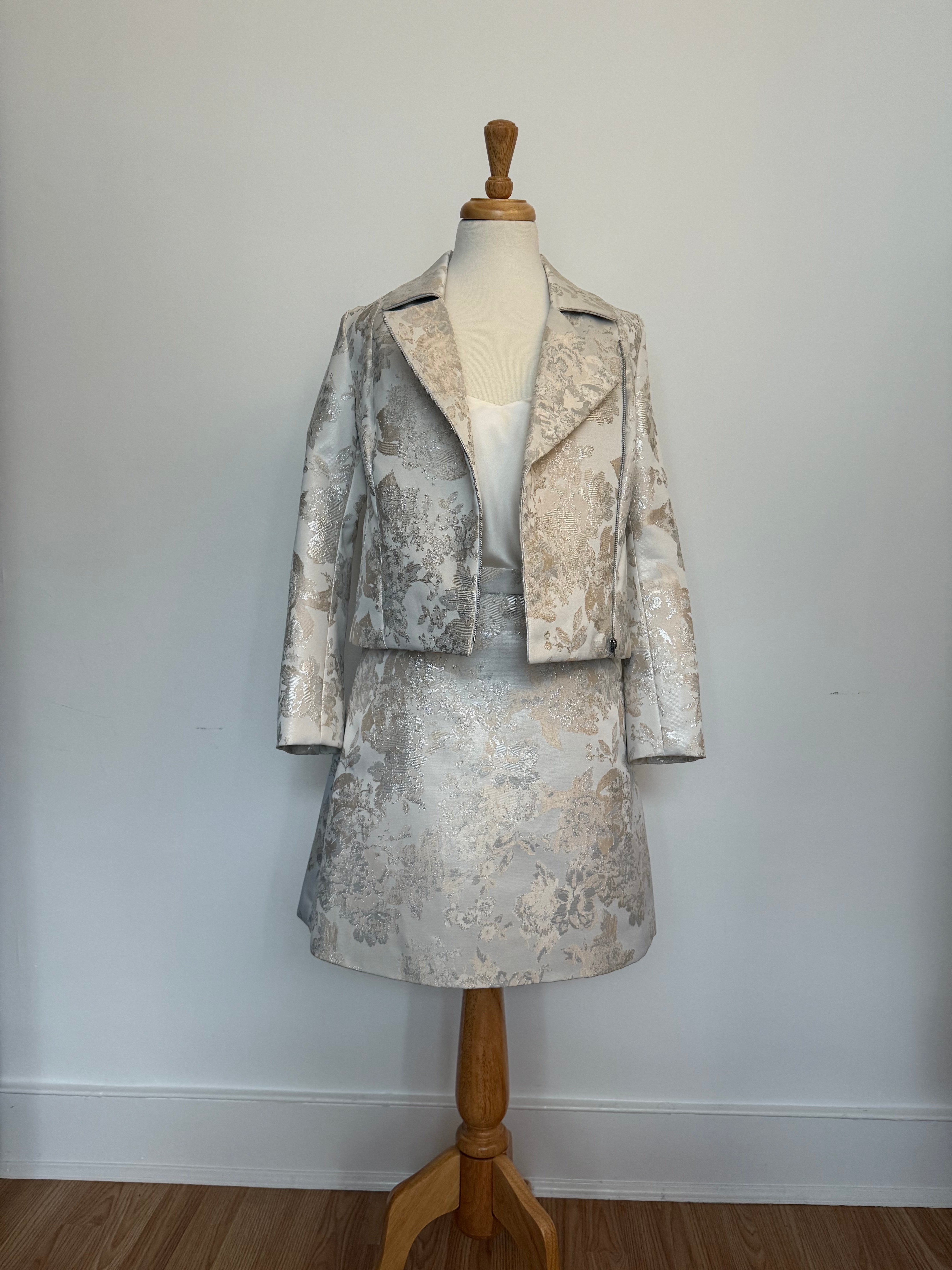 Elegant women's two-piece jacquard outfit on a dress mannequin, featuring a light cream and silver floral-patterned cropped jacket and matching A-line skirt, styled with a soft satin camisole underneath against a plain background.