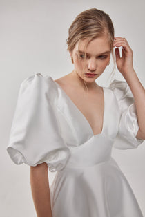A woman wearing a white mini dress with dramatic puff sleeves, a wide plunge V-neckline, and a mini circle skirt with pockets.