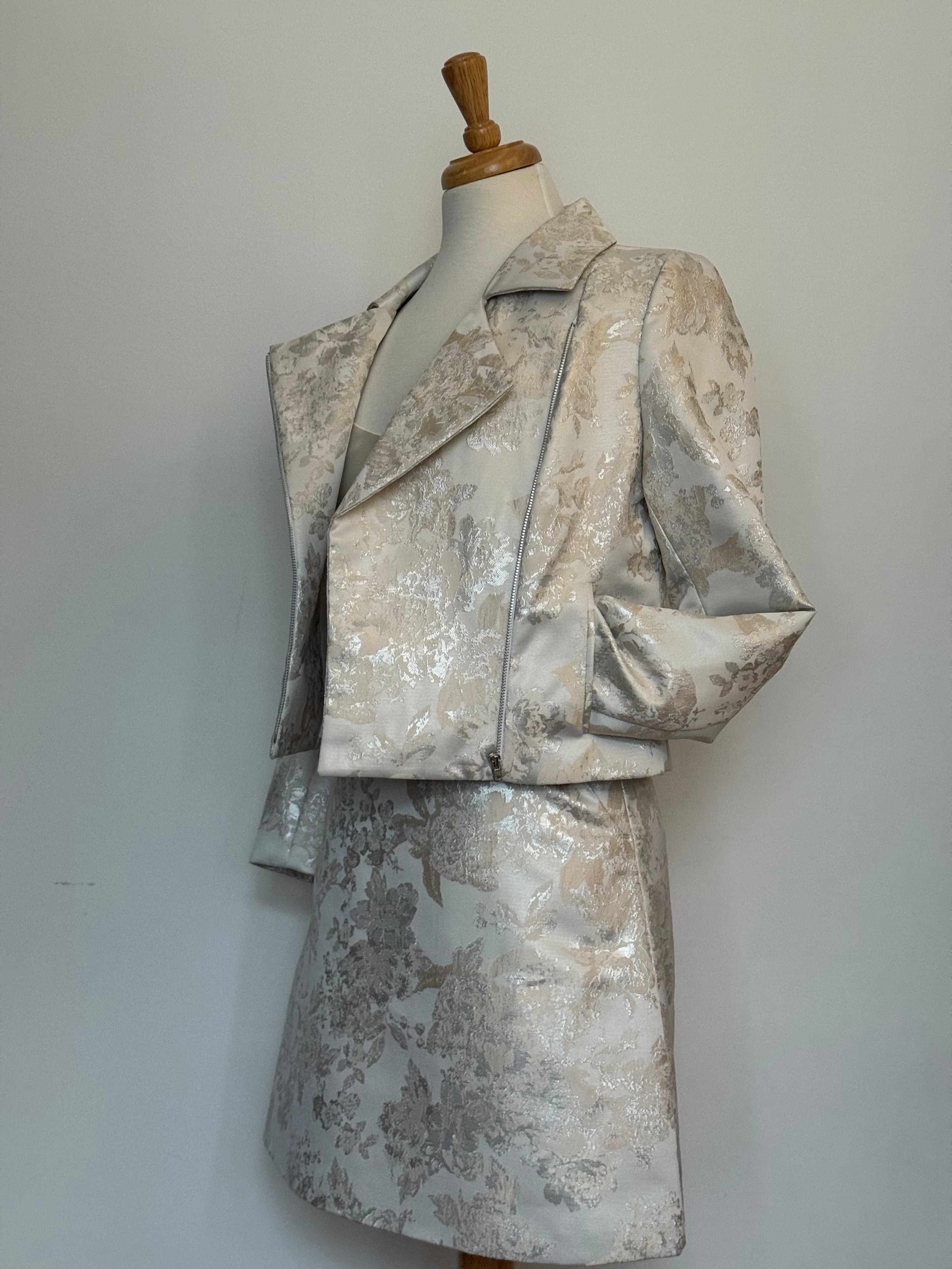 Elegant women's two-piece jacquard outfit on a dress mannequin, featuring a light cream and silver floral-patterned cropped jacket and matching A-line skirt, side styled 