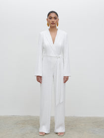 A woman standing wearing a white sequin jumpsuit with long sleeves and a tie at the waist, against a white background.