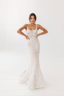 A woman modeling a white, floral lace bridal gown with a square neckline and a slim fit-and-flare silhouette, with a godet train.