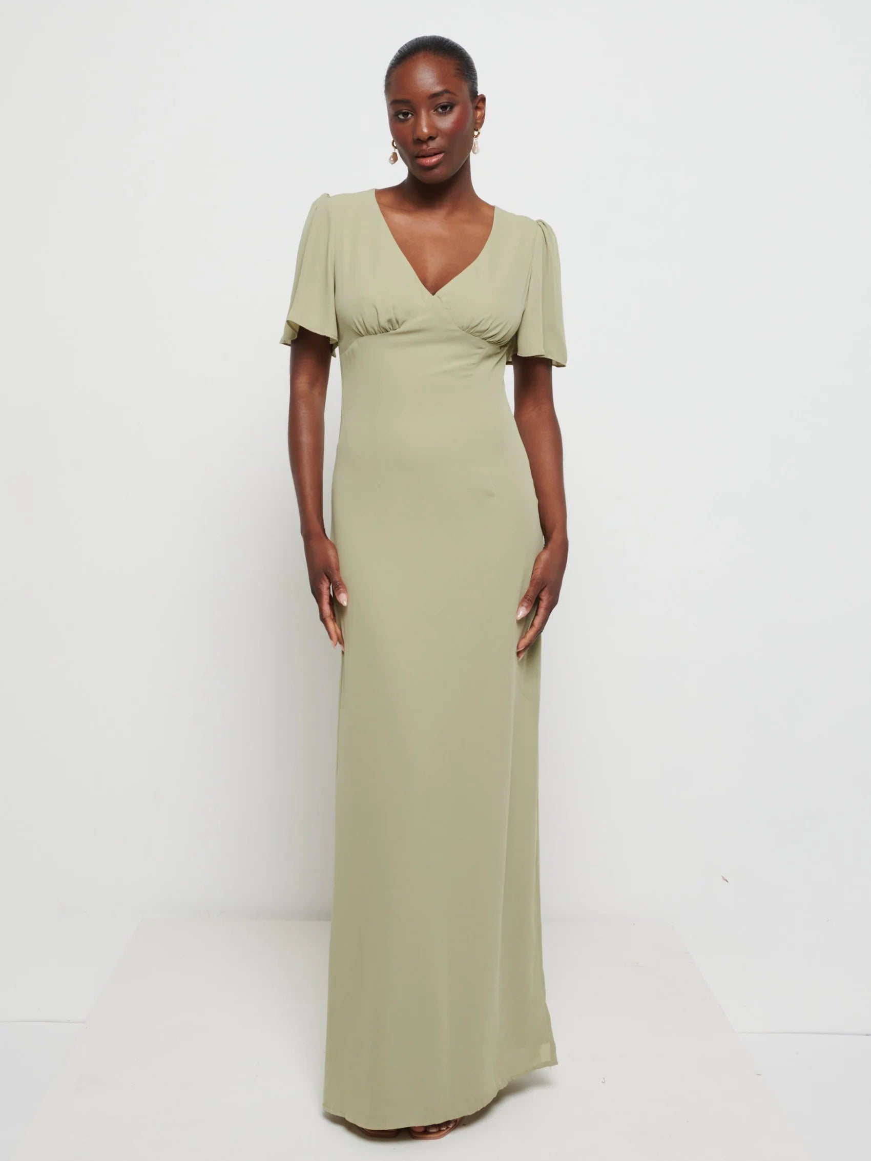 Woman wearing Flattering V-neckline Edie Chiffon Maxi bridesmaid dress in olive green,  under Bust detailing with floaty cap sleeves against a white background
