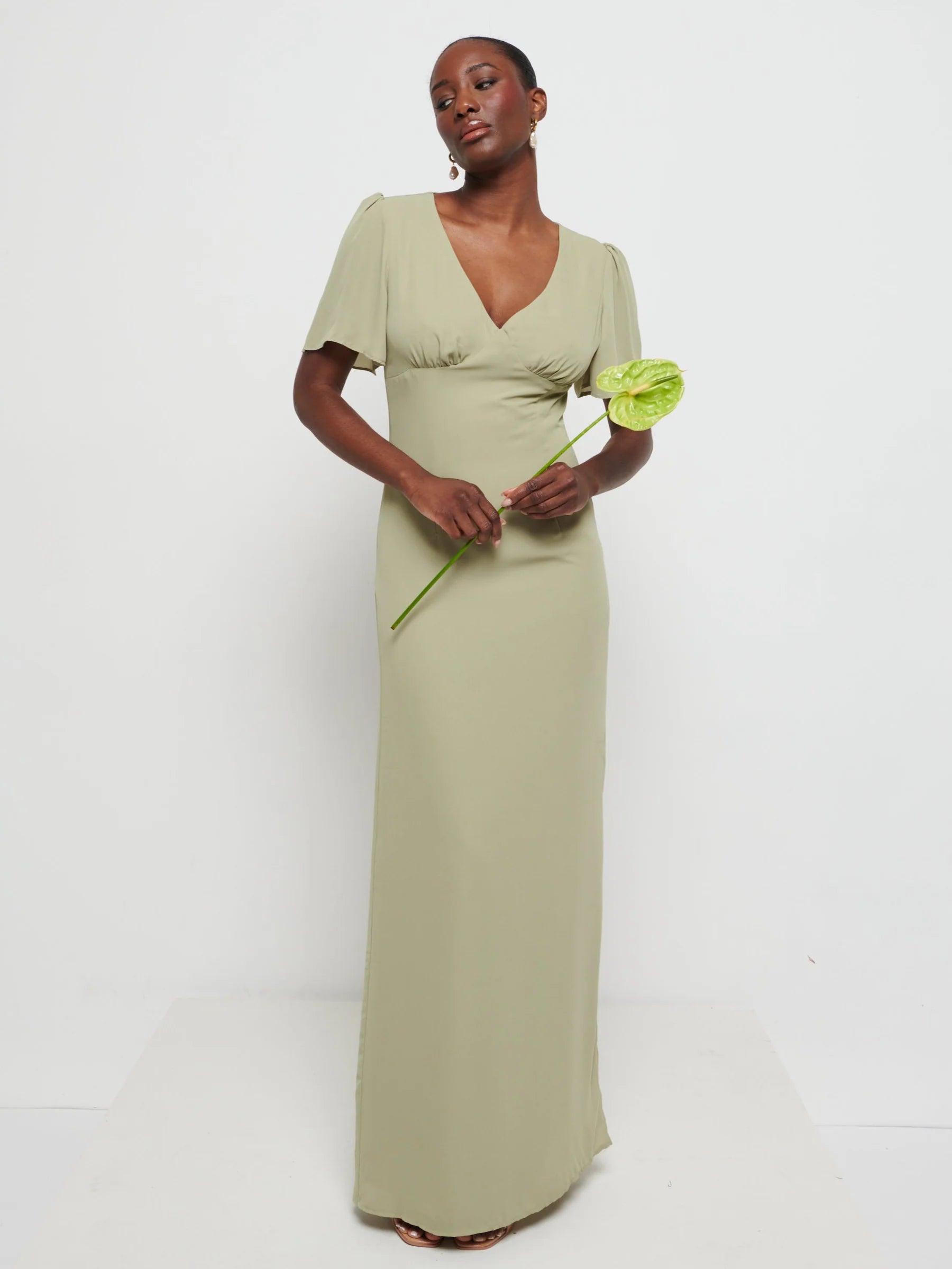 Model in Flattering V-neckline Edie Chiffon Maxi bridesmaid dress in olive green,  under Bust detailing with floaty cap sleeves holding a flower against a white background