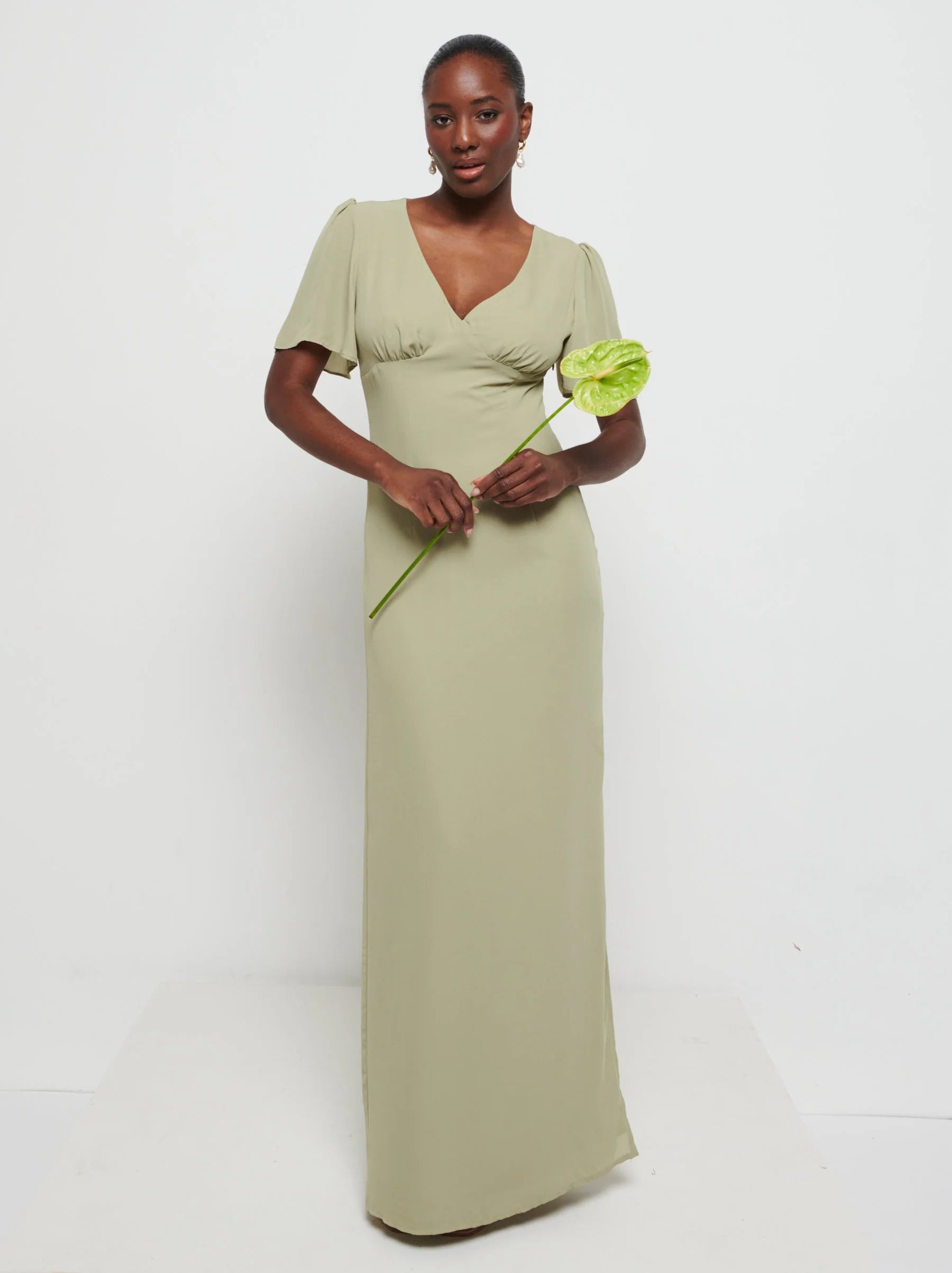 Model in Flattering V-neckline Edie Chiffon Maxi bridesmaid dress in olive green,  under Bust detailing with floaty cap sleeves holding a flower against a white background