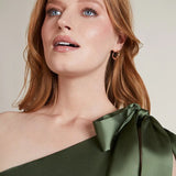 A studio photo of a brunette white woman wearing a body-hugging emerald green satin dress.