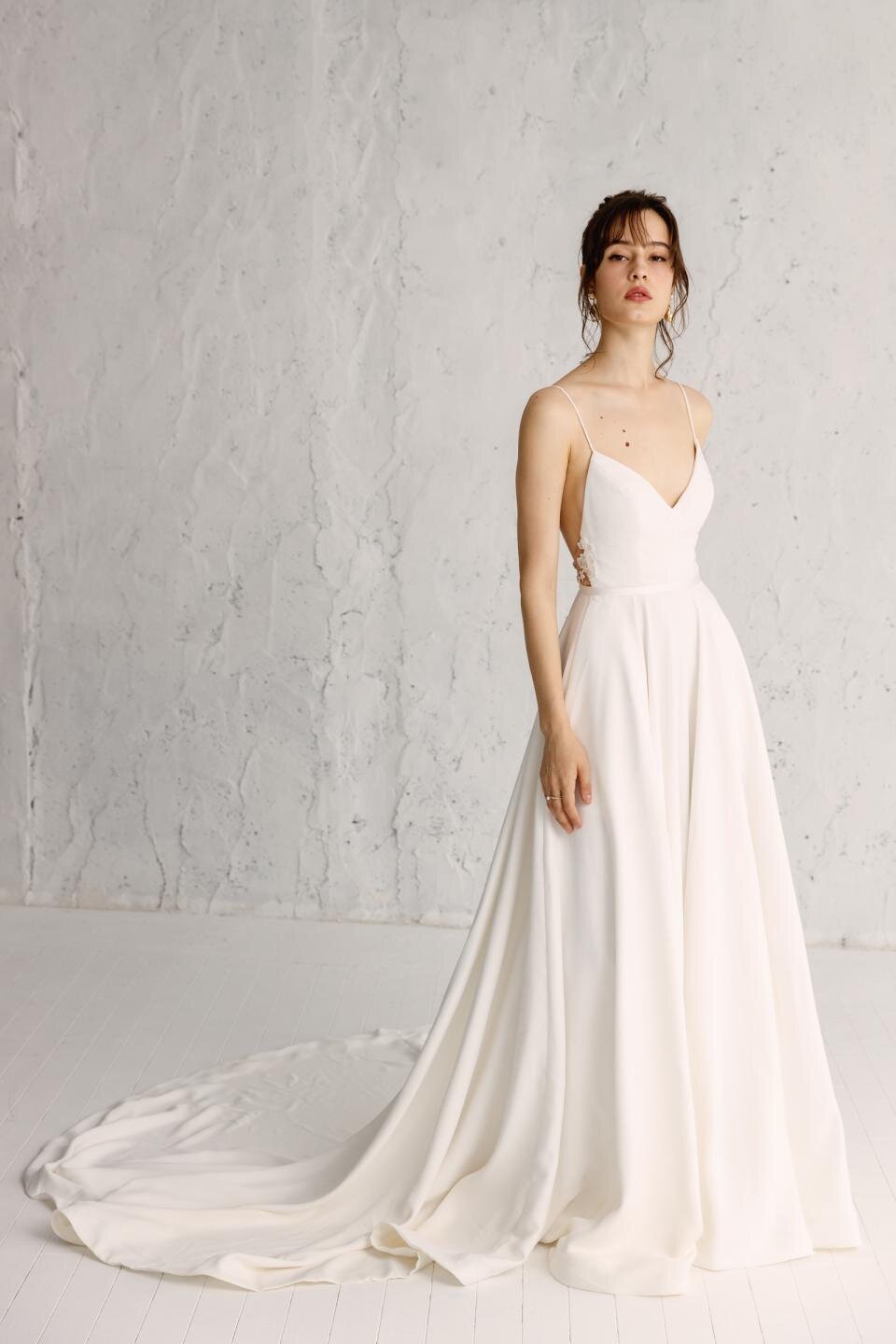 A woman wearing a white full gown with spaghetti straps, hand-appliquéd embroidery, and a cinched waist.