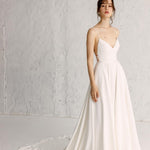 A woman wearing a white full gown with spaghetti straps, hand-appliquéd embroidery, and a cinched waist.