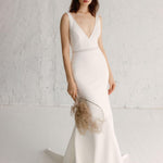 A woman in a white, V-neck, sleeveless, maxi dress with hand-beaded embellishments and a bold, embroidered waistband, standing against a white background.