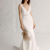 A woman in a white, V-neck, sleeveless, maxi dress with hand-beaded embellishments and a bold, embroidered waistband, standing against a white background.