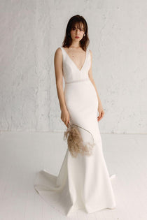A woman in a white, V-neck, sleeveless, maxi dress with hand-beaded embellishments and a bold, embroidered waistband, standing against a white background.