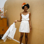 A woman wearing a white satin camisole top with a floral headband and holding a white jacket.