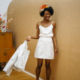 A woman wearing a white satin camisole top with a floral headband and holding a white jacket.