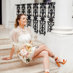 A woman sitting on steps wearing a white short bridal dress with embroidered polka dots, puff sleeves, and a mini skirt length. She has orange heels and is holding a bouquet.