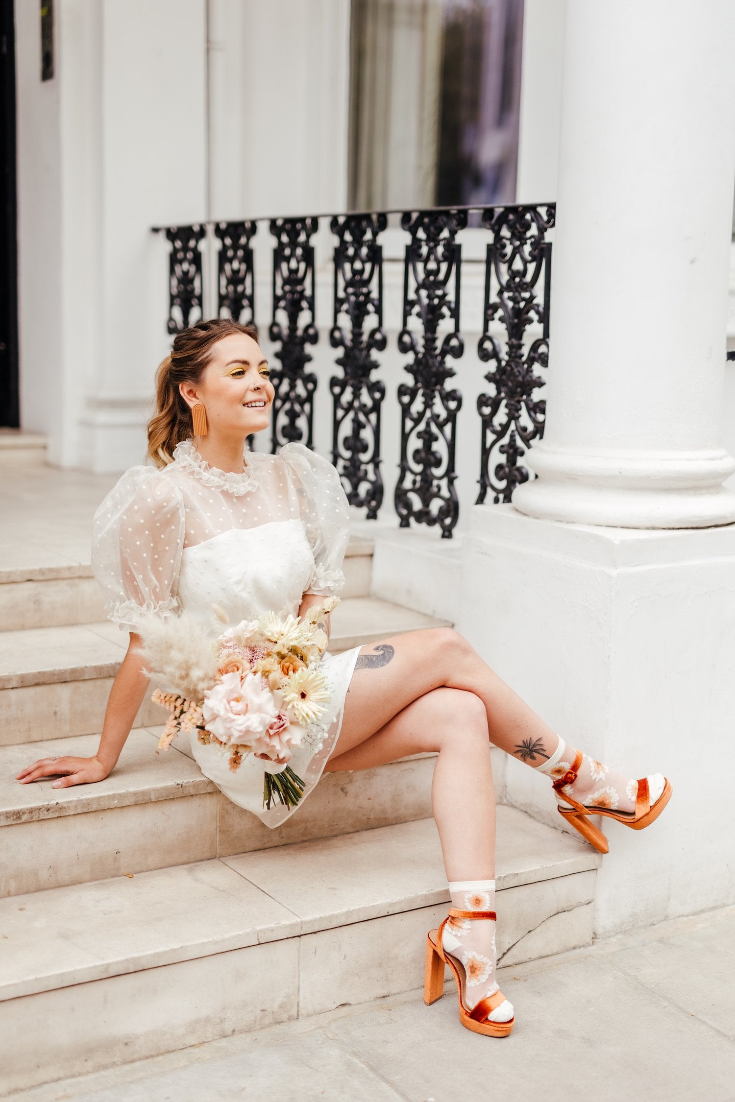 A woman sitting on steps wearing a white short bridal dress with embroidered polka dots, puff sleeves, and a mini skirt length. She has orange heels and is holding a bouquet.