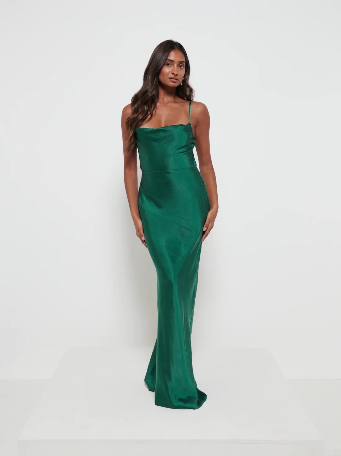 A woman wearing an emerald green maxi dress with a cowl neckline and spaghetti straps, made from a luxurious satin fabric.