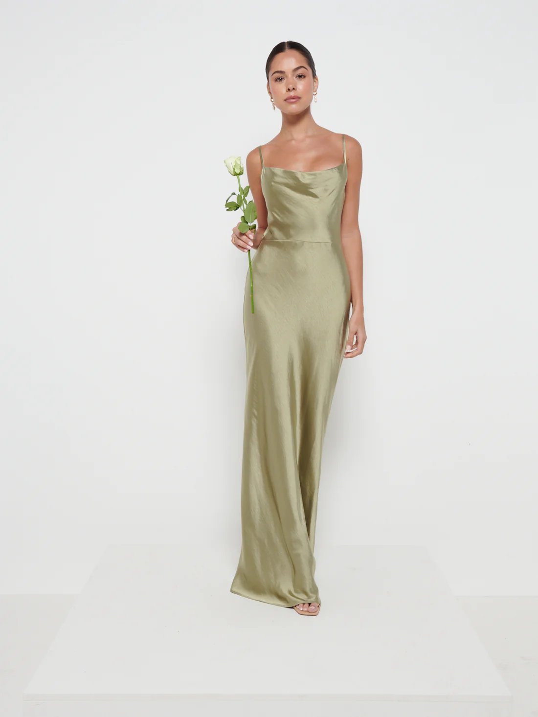 A woman wearing an olive green maxi dress with a cowl neckline and spaghetti straps, holding a small bouquet.