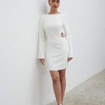 A woman standing against a white background, wearing a white mini dress with a high neckline, wide kimono sleeves, and a concealed zipper at the back. She is also wearing white open-toe heeled sandals.