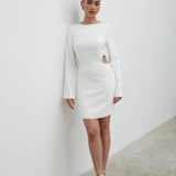 A woman standing against a white background, wearing a white mini dress with a high neckline, wide kimono sleeves, and a concealed zipper at the back. She is also wearing white open-toe heeled sandals.