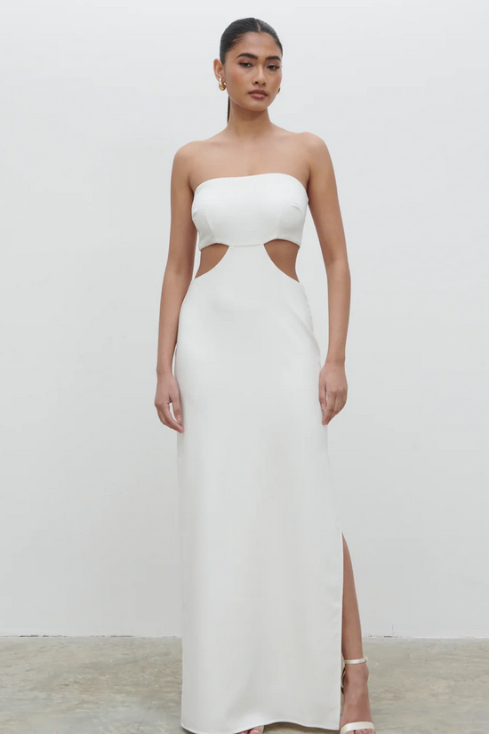 A woman wearing a white maxi dress with a bandeau neckline, cut-out designs, and a side slit. She is posing against a plain background.