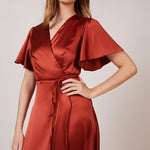A woman wearing a burnt orange satin bridesmaid dress with a v-neck, short draped sleeves, and a waterfall hem.