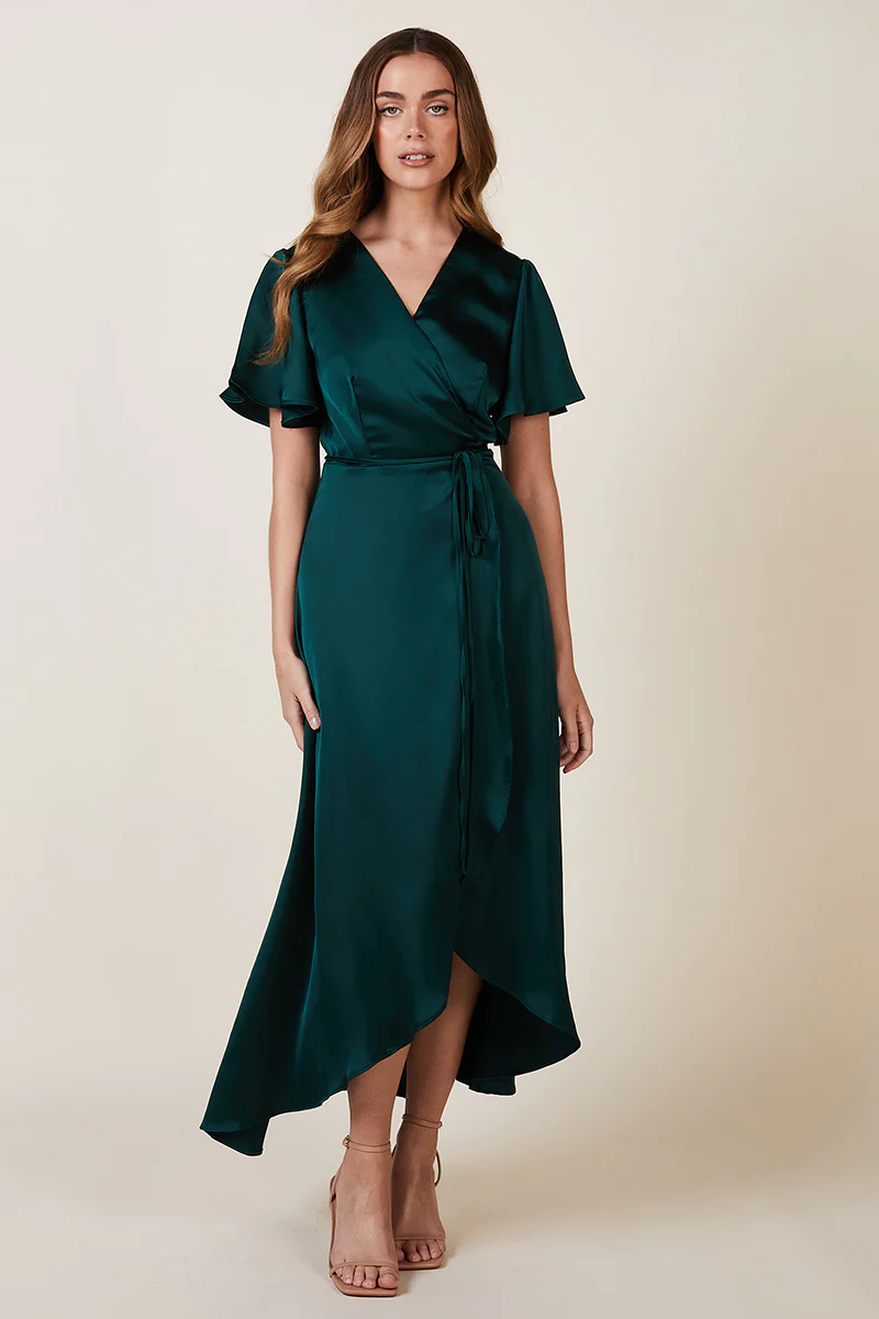 A Model wearing a forest green satin  dress with flutter sleeves and a tie waist, standing a, paired with nude high-heeled sandals.