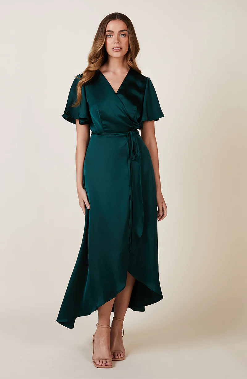 A Model wearing a forest green satin  dress with flutter sleeves and a tie waist, standing a, paired with nude high-heeled sandals.