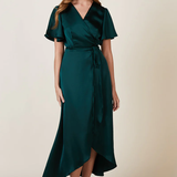 A Model wearing a forest green satin  dress with flutter sleeves and a tie waist, standing a, paired with nude high-heeled sandals.