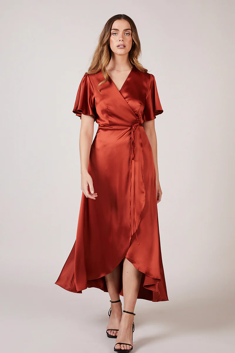 A woman wearing a burnt orange satin bridesmaid dress with a v-neck, short draped sleeves, and a waterfall hem.