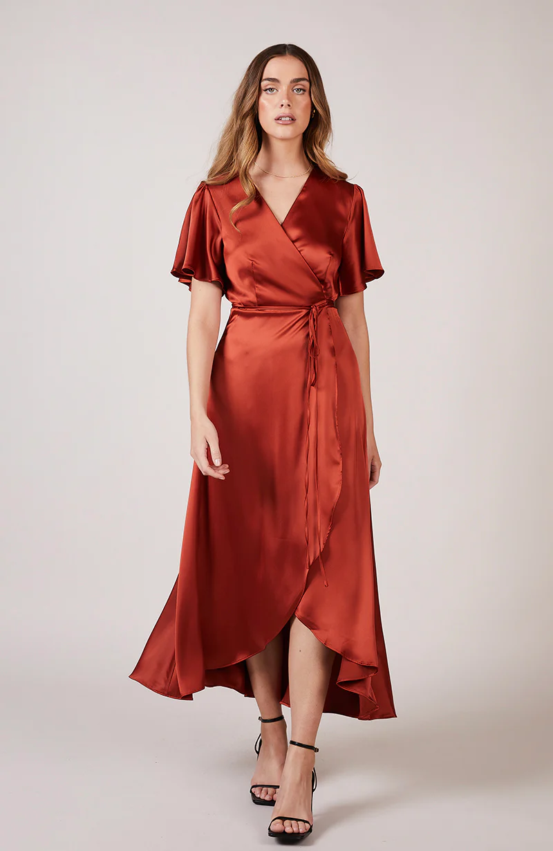 A woman wearing a burnt orange satin bridesmaid dress with a v-neck, short draped sleeves, and a waterfall hem.