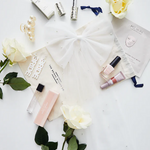 An assortment of bridal beauty products including skincare, makeup, and accessories spread out on a surface, with a mini tulle veil and bridal accessories like earrings and a bow included.
