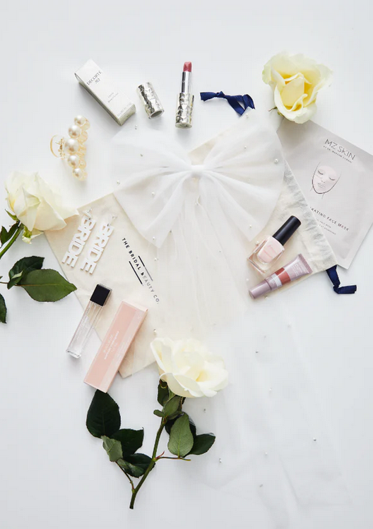 An assortment of bridal beauty products including skincare, makeup, and accessories spread out on a surface, with a mini tulle veil and bridal accessories like earrings and a bow included.