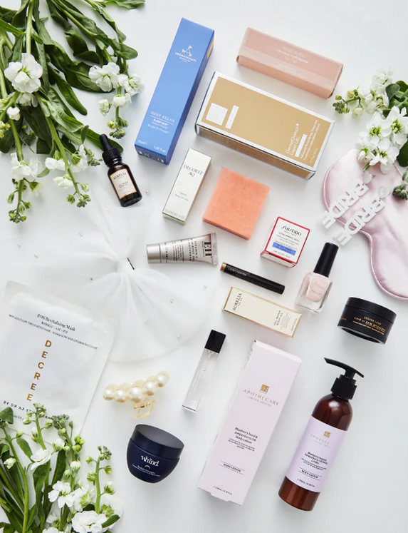 An assortment of luxury beauty products from The Ultimate Bridal Beauty Box, including skincare, cosmetics, and accessories displayed on a white surface with floral decorations.