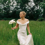 A woman in an off-the-shoulder white wedding dress with corset detail, holding a bouquet, standing in a grassy field.