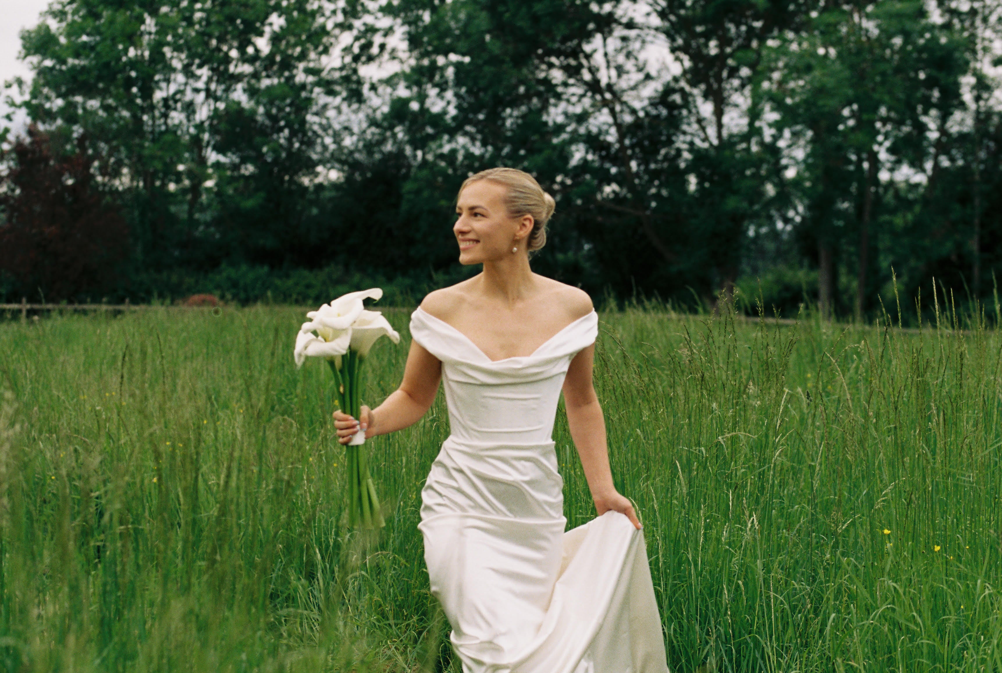 A woman in an off-the-shoulder white wedding dress with corset detail, holding a bouquet, standing in a grassy field.