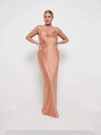 A woman wearing a cinnamon-colored maxi dress with a cowl neckline and spaghetti straps, floor-length, made from satin fabric.
